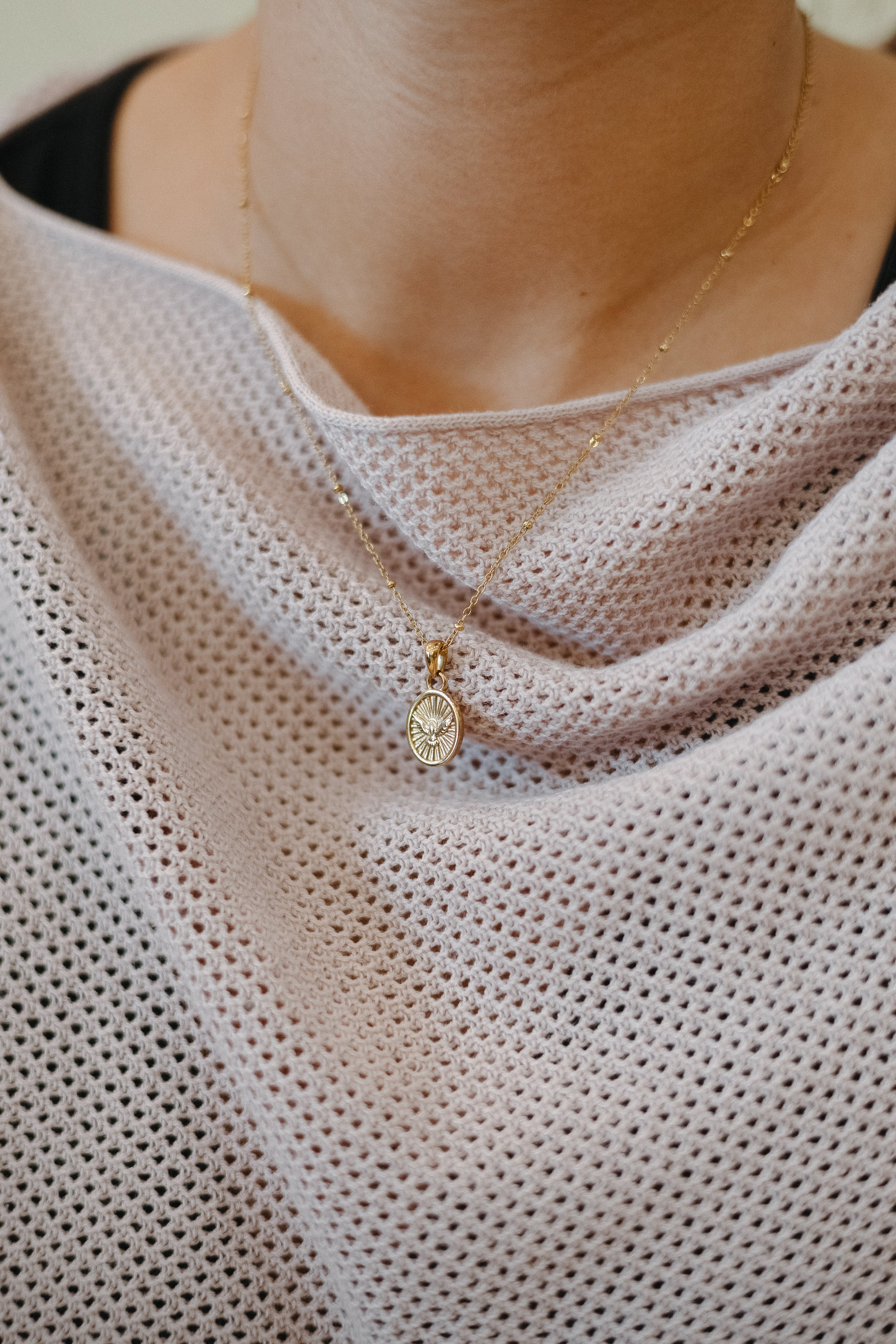 Close up of a woman wearing a delicate gold Holy Spirit necklace with an oval dove pendant over a white knit top by Blessed Is She