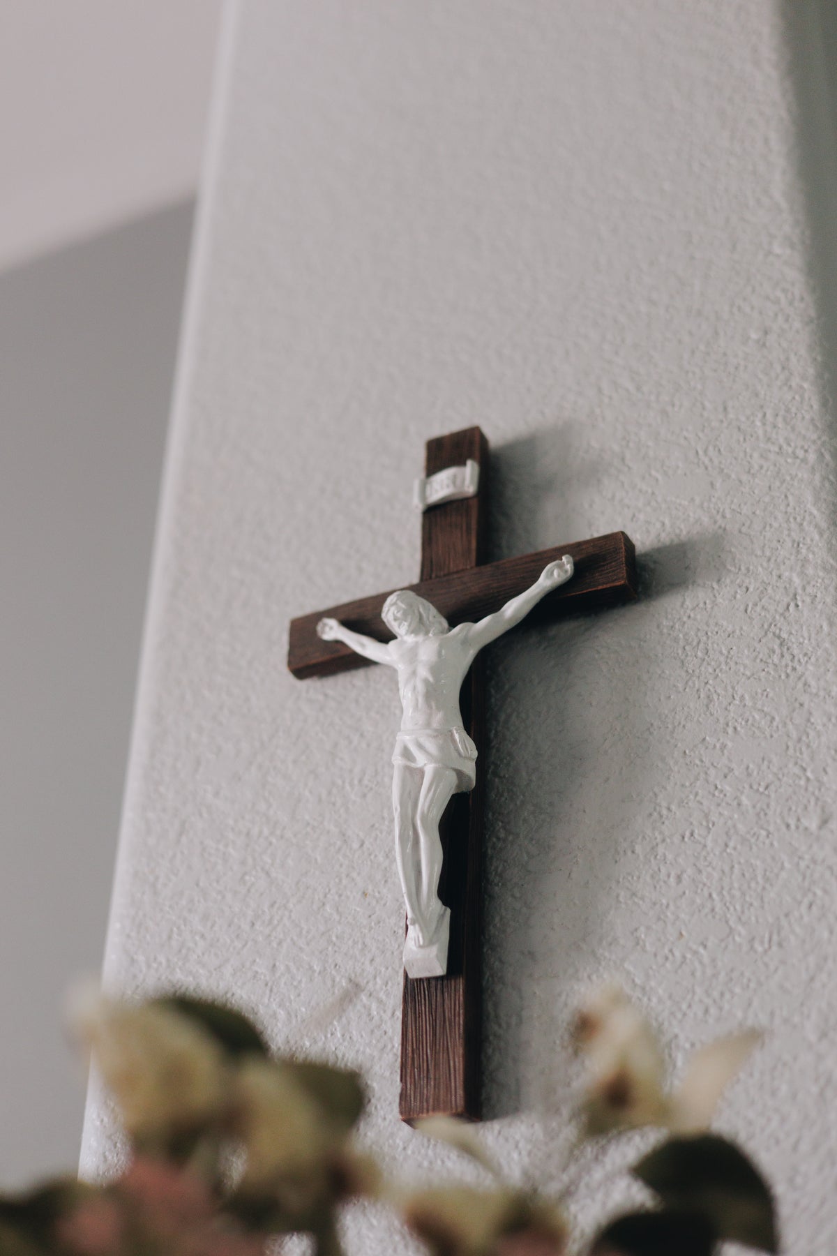 Dark brown wall crucifix with a white corpus of Christ mounted on a textured wall, softly lit above blurred flowers in a peaceful Catholic setting