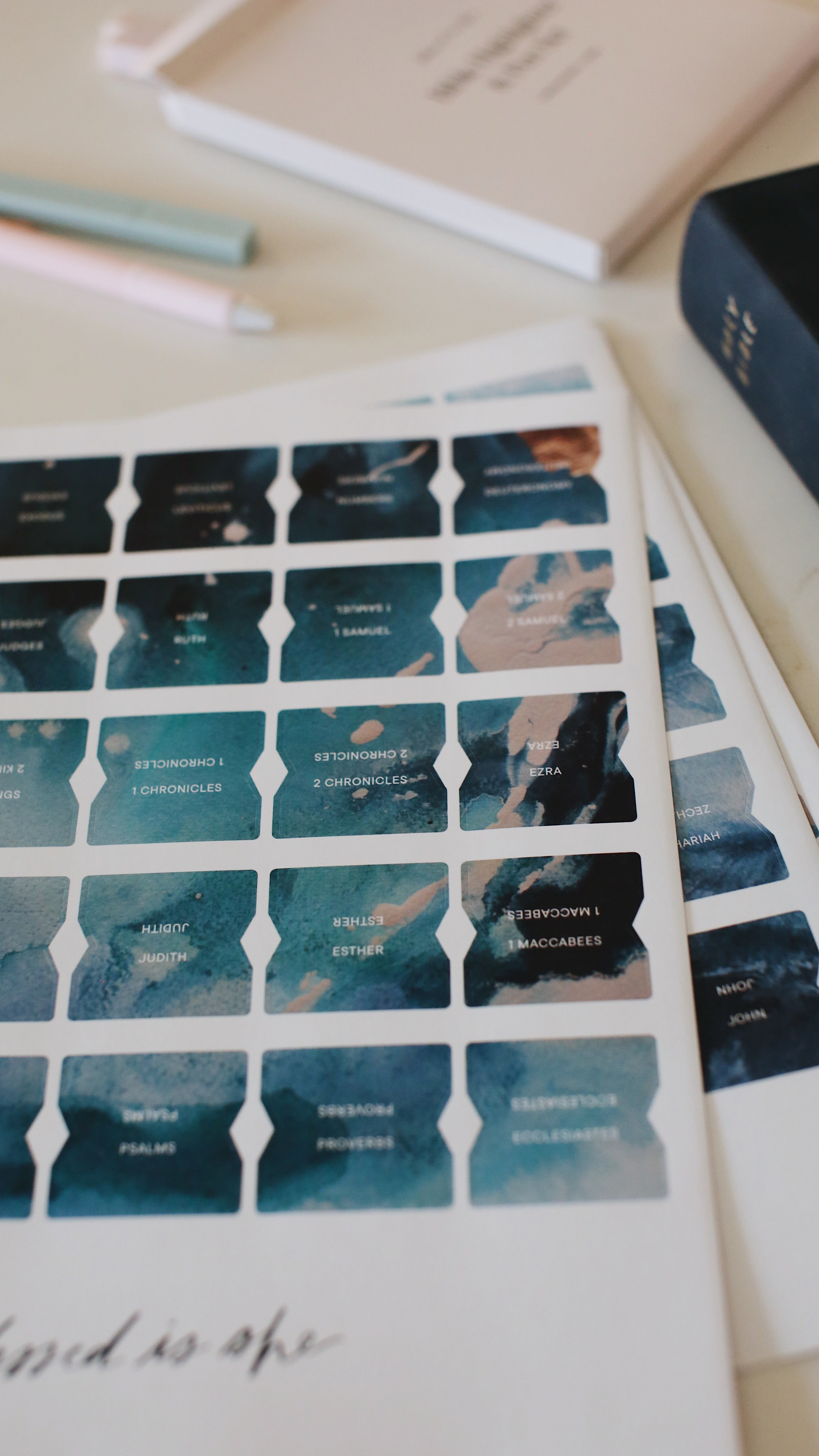 Sheet of teal and navy Bible study tabs for women beside a book and pastel pens, designed to organize Scripture for peaceful daily prayer.